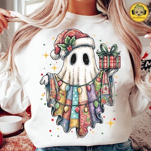 Puede incluir: Sudadera blanca con un fantasma de dibujos animados que lleva un gorro de Papá Noel y sostiene un regalo envuelto. El fantasma está cubierto con un edredón de retazos colorido con luces navideñas. Estrellas y puntos festivos rodean al fantasma.