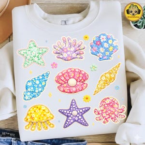 May include: White sweatshirt featuring colorful seashell and starfish designs. The designs include a green starfish, a purple seashell, a blue seashell, a pink clam shell, a yellow jellyfish, and a purple starfish. The designs are decorated with floral patterns.