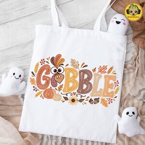 May include: A white tote bag featuring the text GOBBLE in large, orange and brown striped letters. The letter O is replaced by a turkey face. The design includes autumn leaves, pumpkins, sunflowers, and acorns in shades of orange and brown.
