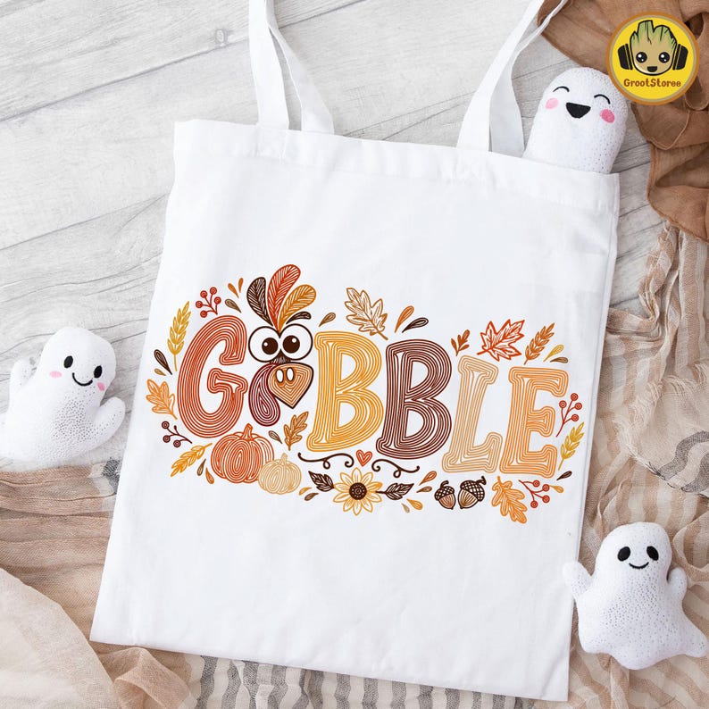 May include: A white tote bag featuring the text GOBBLE in large, orange and brown striped letters. The letter O is replaced by a turkey face. The design includes autumn leaves, pumpkins, sunflowers, and acorns in shades of orange and brown.