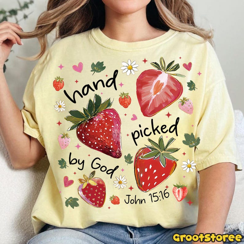 May include: A pale yellow t-shirt with a strawberry design and the text "hand picked by God" and John 15:16. The design includes strawberries, flowers, and hearts. The shirt has a crew neck and short sleeves.