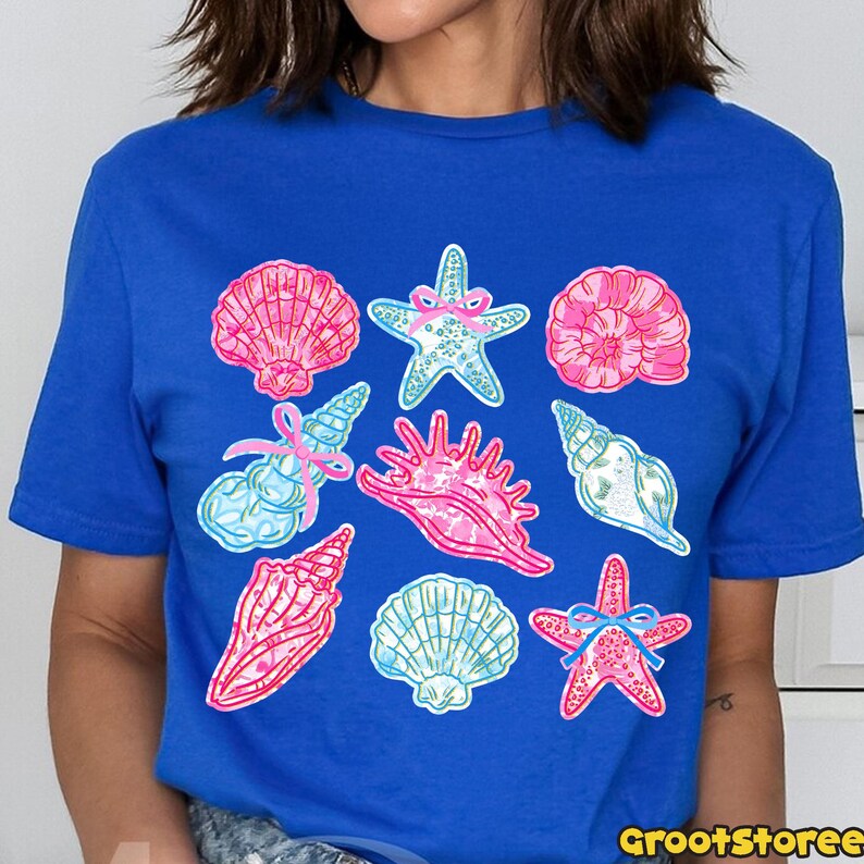 May include: A royal blue t-shirt featuring a collection of colorful seashell and starfish illustrations. The designs include pink, blue, and white shells and starfish with bow accents. The shirt has a crew neck and short sleeves.