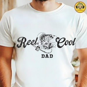 Può includere: T-shirt bianca con grafica nera raffigurante un pesce che salta fuori dall'acqua, una canna da pesca e la scritta "Reel Cool Dad". Il design è in stile vintage.