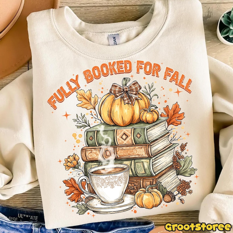 May include: Cream sweatshirt with the text "FULLY BOOKED FOR FALL" in orange. The graphic features a stack of books, pumpkins, autumn leaves, and a cup of coffee. A cozy design for autumn and book lovers.