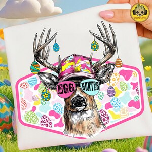 Op de afbeelding: Wit T-shirt met een hert met een zonnebril met de tekst "EGG HUNTER" en een kleurrijke hoed. Het ontwerp bevat paaseieren en een roze rand. Paas-thema kleding.