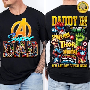 Puede incluir: Camiseta negra con un gráfico colorido de "Super Papá" con personajes de superhéroes. El diseño frontal incluye la palabra "Super" en azul y amarillo, y "DAD" lleno de arte de cómic. La parte trasera dice "Papá, eres mi superhéroe".