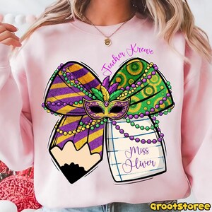 May include: A light pink sweatshirt featuring a Mardi Gras-themed design. The design includes a bow with purple and gold stripes, green and gold polka dots, a mask, and a pencil. The text "Teacher Krewe" and "Miss Oliver" are also present.