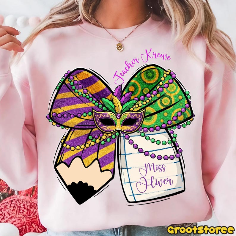 May include: A light pink sweatshirt featuring a Mardi Gras-themed design. The design includes a bow with purple and gold stripes, green and gold polka dots, a mask, and a pencil. The text "Teacher Krewe" and "Miss Oliver" are also present.