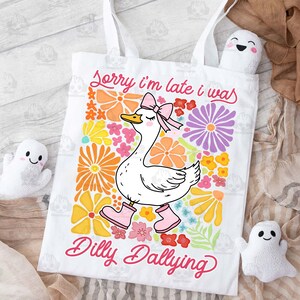 Op de afbeelding: Witte tote bag met een roze en witte gans illustratie en de tekst "Sorry, ik ben te laat, ik was aan het dralen". De gans draagt roze schoenen en is omringd door kleurrijke bloemen.