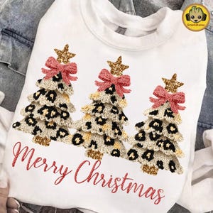 May include: White sweatshirt with three leopard print Christmas tree designs. Each tree has a pink bow, a gold star, and a gold glitter base. The words "Merry Christmas" are embroidered in red.