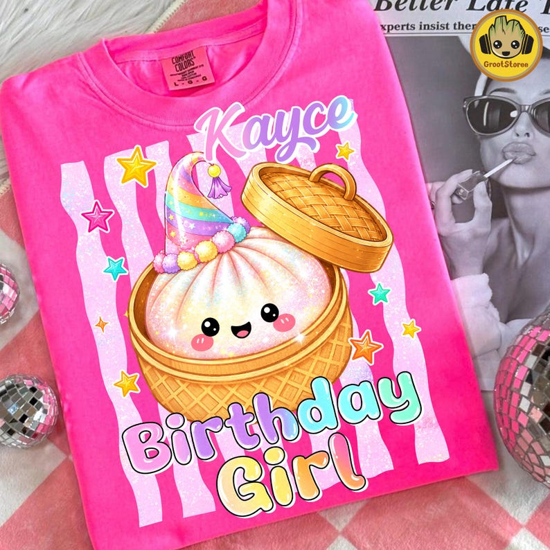 May include: A bright pink t-shirt featuring a graphic of a smiling bao bun in a bamboo steamer basket. The bun wears a rainbow party hat. The text reads Kayce Birthday Girl in colourful letters, surrounded by small stars and light pink vertical stripes.