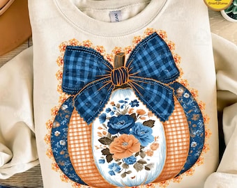 May include: Cream-coloured sweatshirt with a pumpkin design. The pumpkin has a blue plaid bow, orange and blue plaid sections, and a floral arrangement of blue and orange flowers. The design is surrounded by an orange lace trim.