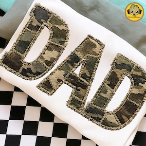 May include: White pillow with the word "DAD" in large, block letters. The letters are made of camouflage fabric with a stitched, frayed edge. The pillow is on a black and white checkered surface.