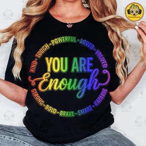 Pode incluir: Camiseta preta com um design de arco-íris que diz "YOU ARE Enough". O design é rodeado por palavras como "Kind", "Strong" e "Brave". A camiseta tem gola redonda e mangas curtas.