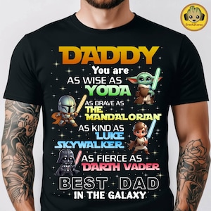 Puede incluir: Camiseta negra con el texto "DADDY" y un tema de Star Wars. La camiseta presenta gráficos de Yoda, The Mandalorian, Luke Skywalker y Darth Vader, con las palabras "Best Dad in the Galaxy".