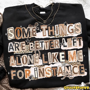 Puede incluir: Sudadera negra con el texto "SOME THINGS ARE BETTER LEFT ALONE LIKE ME FOR INSTANCE" en un estilo collage con detalles de estampado de leopardo, lazos y estrellas. Un collar dorado est&aacute; sobre la sudadera.