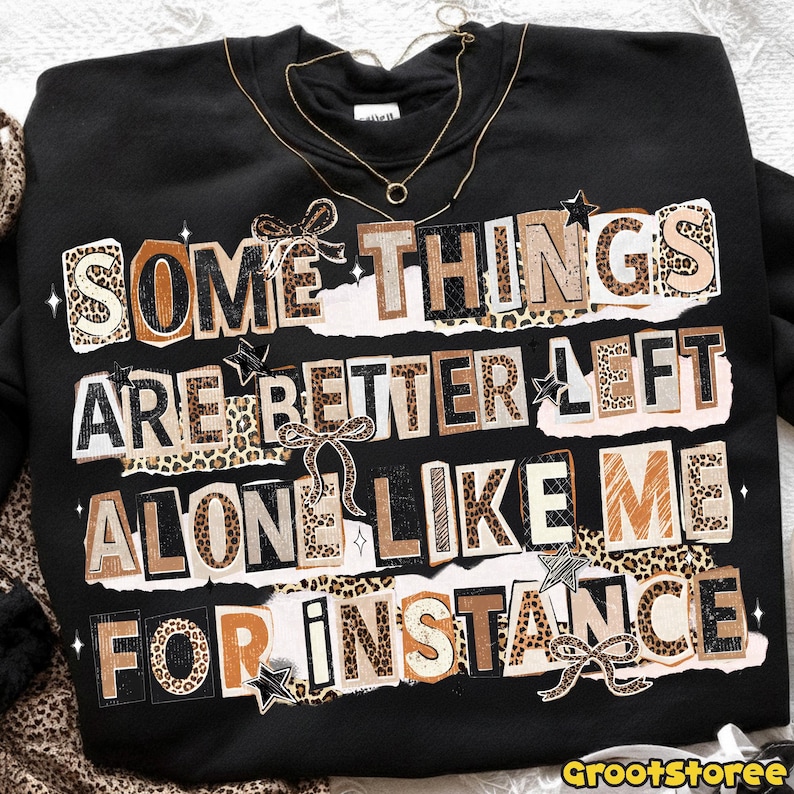 Puede incluir: Sudadera negra con el texto "SOME THINGS ARE BETTER LEFT ALONE LIKE ME FOR INSTANCE" en un estilo collage con detalles de estampado de leopardo, lazos y estrellas. Un collar dorado est&aacute; sobre la sudadera.