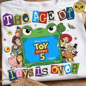 May include: A white t-shirt featuring a graphic of Toy Story characters surrounding a green frog-shaped frame. The text reads THE AGE OF TOY STORY 5 ONLY IN THEATERS JUNE 19 TOYS IS OVER. The design includes Buzz Lightyear, Woody, Jessie, and other characters.
