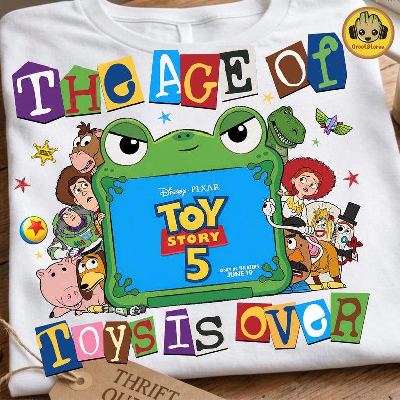 May include: A white t-shirt featuring a graphic of Toy Story characters surrounding a green frog-shaped frame. The text reads THE AGE OF TOY STORY 5 ONLY IN THEATERS JUNE 19 TOYS IS OVER. The design includes Buzz Lightyear, Woody, Jessie, and other characters.