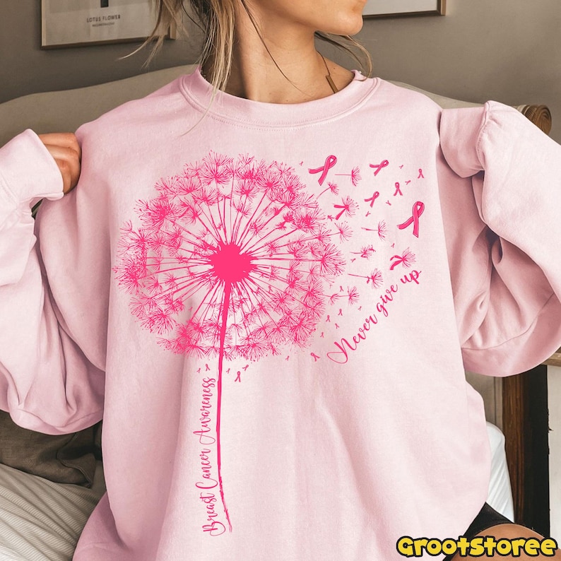May include: A light pink sweatshirt featuring a large pink dandelion design with the words "Breast Cancer Awareness" and "Never give up". Pink ribbons are scattered around the dandelion. The sweatshirt is a soft, comfortable material.