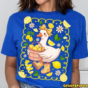 May include: A blue t-shirt features a cartoon goose with a yellow bow, holding a basket of lemons. The design includes lemons, daisies, and lemon peel accents within a yellow border. The text "GrootStoree" is at the bottom.