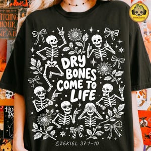 Könnte beinhalten: Ein schwarzes T-Shirt mit einem weißen Grafikdruck von fünf Skeletten in verschiedenen Posen, umgeben von Blumen, Schleifen und Sternen. Der Text in der Mitte lautet "DRY BONES COME TO LIFE" mit "EZEKIEL 37:1-10" in Weiß am unteren Rand.