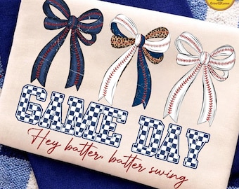May include: Cream-colored t-shirt featuring three decorative bows with baseball stitching and a leopard print accent. The words "GAME DAY" are in a checkered pattern, with "Hey batter, batter swing" in red script below.