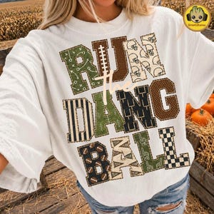 May include: A white crewneck t-shirt featuring the text RUN THE DANG BALL in large block letters. Each letter has a unique pattern including football plays, helmets, stripes, footballs, pennants, foam fingers, and a black and white checkerboard.