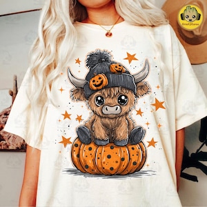 Op de afbeelding: Crèmekleurig T-shirt met een cartoon Highland-koe met een grijze beanie met pompoenaccenten, zittend op een oranje pompoen. Het ontwerp bevat oranje sterren, wat een Halloween-thema creëert.