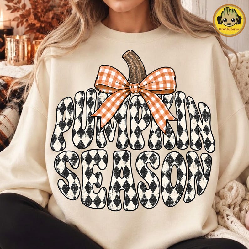 May include: A beige crewneck sweatshirt featuring a large graphic of a pumpkin with a black and white diamond pattern. The pumpkin is topped with an orange and white gingham bow and the text PUMPKIN SEASON in a bold, wavy black font.