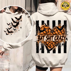 May include: A cream-colored hooded sweatshirt featuring a large graphic on the back. The design includes a heart shape filled with orange spiderwebs, black bats, and spiders over vertical black stripes, with the text BAT SHIT CRAZY printed across.