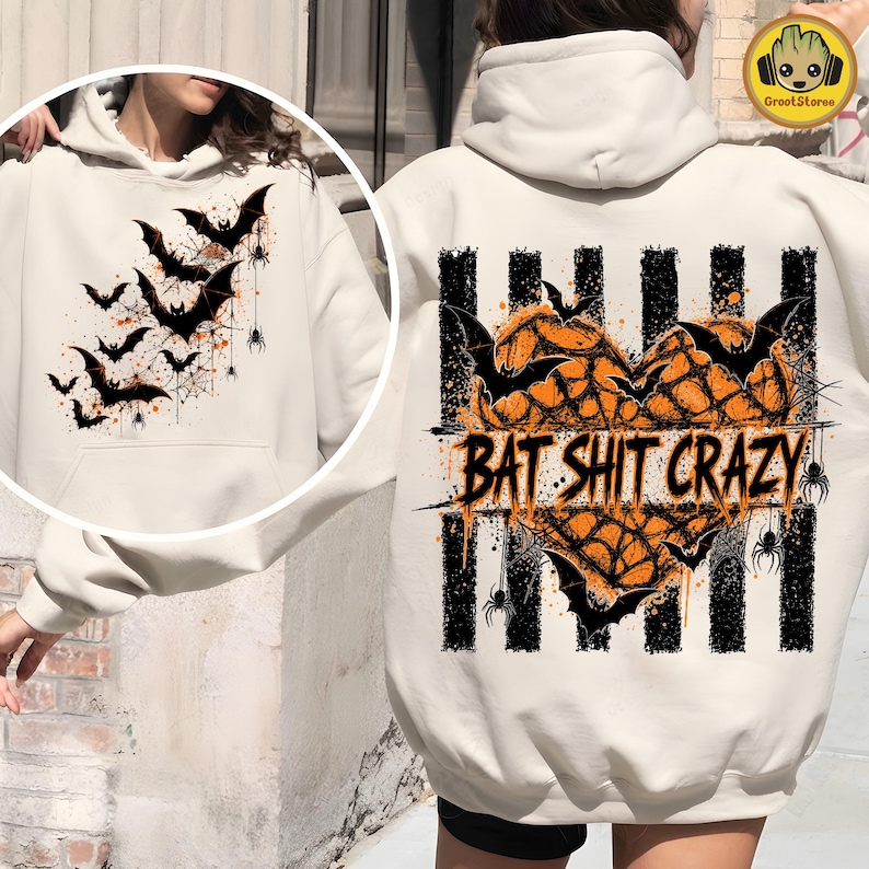 May include: A cream-colored hooded sweatshirt featuring a large graphic on the back. The design includes a heart shape filled with orange spiderwebs, black bats, and spiders over vertical black stripes, with the text BAT SHIT CRAZY printed across.