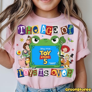 May include: A light pink t-shirt featuring a graphic print of a green frog-shaped frame containing the text Disney Pixar Toy Story 5. The design includes various animated characters surrounding the frame and the text The Age Of Toys Is Over.
