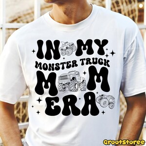 K&ouml;nnte beinhalten: Wei&szlig;es T-Shirt mit schwarzem Grafikdesign. Das Design zeigt den Text "IN MY MONSTER TRUCK MOM ERA" in einer fetten, retro Schriftart, zusammen mit Illustrationen von Monstertrucks und Sternen. Das Shirt ist ein l&auml;ssiger Alltagsartikel.