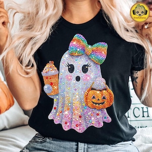 May include: Black t-shirt featuring a rhinestone ghost with a rainbow bow, holding a pumpkin and a drink. The ghost has large eyes and a smiling mouth. The shirt is a Halloween-themed design.