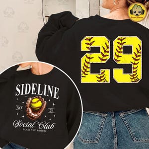 May include: Black sweatshirt with a softball design on the back. The design features the number 29 with a yellow background and red stitching. The front of the sweatshirt has a white graphic with the text "Sideline Social Club" and a baseball glove with a ball.