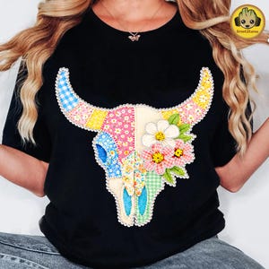 Könnte beinhalten: Schwarzes T-Shirt mit einem Patchwork-Design eines Bullenschädels. Der Schädel ist mit floralen und Vichy-Mustern in Rosa, Blau, Gelb und Grün verziert. Weiße Blumen und grüne Blätter ergänzen das Design.