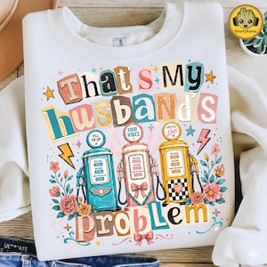 Pode incluir: Camisola branca com a frase "That's My Husband's Problem" em letras coloridas. O design inclui três bombas de gasolina vintage, flores, estrelas e raios. A frase "Good Vibes" também está incluída.