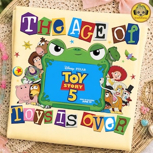 May include: A light yellow t-shirt featuring a graphic of a green frog holding a blue tablet screen that displays the text Disney Pixar Toy Story 5 Only In Theaters June 19. The shirt includes the text The Age Of Toys Is Over and various characters.