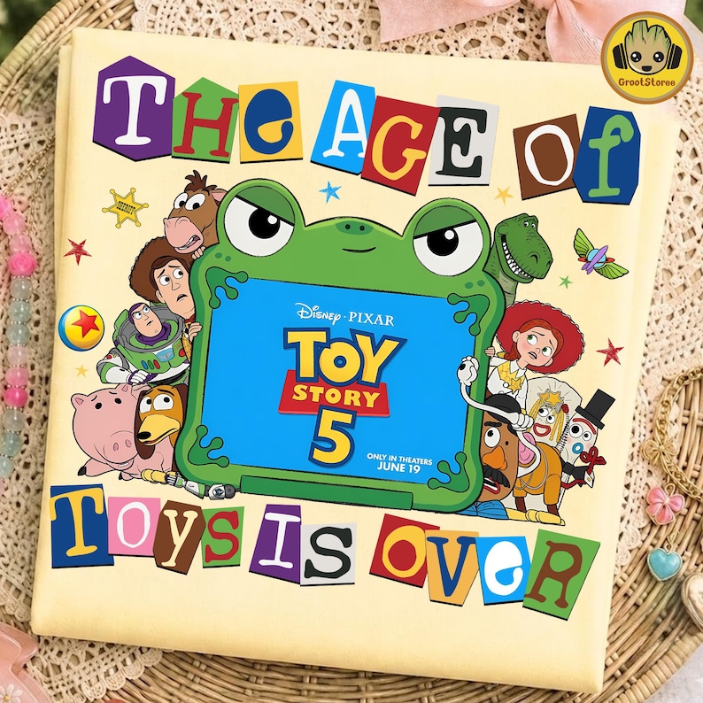 May include: A light yellow t-shirt featuring a graphic of a green frog holding a blue tablet screen that displays the text Disney Pixar Toy Story 5 Only In Theaters June 19. The shirt includes the text The Age Of Toys Is Over and various characters.