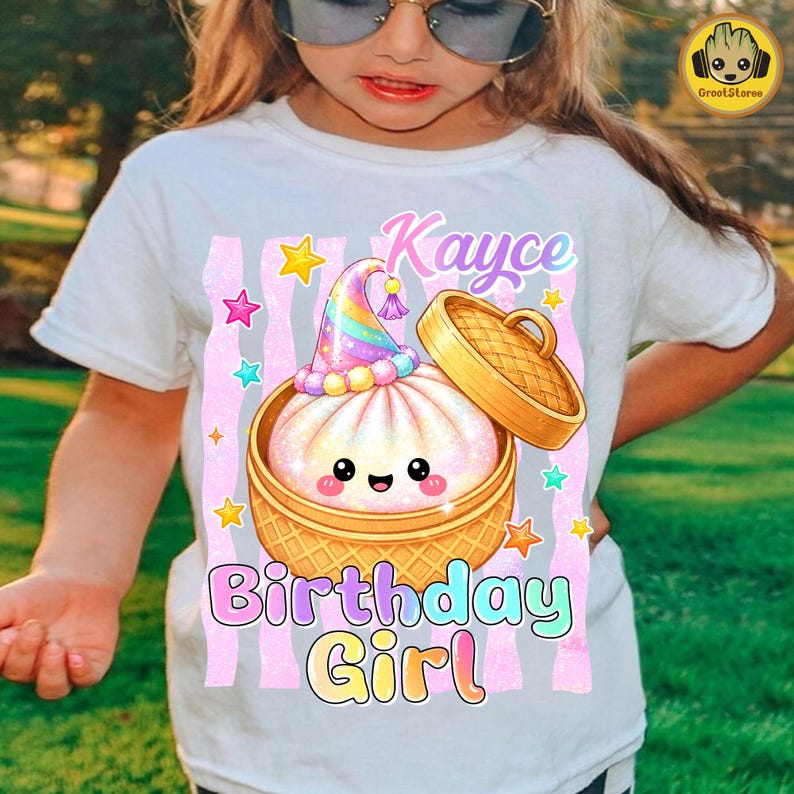May include: A white t-shirt featuring a graphic of a smiling, iridescent dumpling in a bamboo steamer basket, wearing a rainbow party hat. The design includes pink glittery vertical stripes, colourful stars, and the text Kayce Birthday Girl.