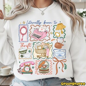 May include: White sweatshirt with a colorful graphic design featuring vintage items like a pink typewriter, mirror, and shopping cart. The text "Literally born to Thrift" is displayed above the images. The design includes a telephone, dishes, and a teapot.