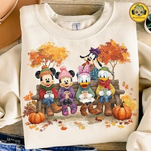 May include: Cream-colored sweatshirt featuring a fall scene with Disney characters. Mickey, Minnie, Daisy, Donald, and Goofy are seated on a bench, holding mugs. Pumpkins and autumn leaves surround them. The sweatshirt has a crew neck.