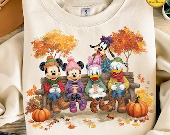 May include: Cream-colored sweatshirt featuring a fall scene with Disney characters. Mickey, Minnie, Daisy, Donald, and Goofy are seated on a bench, holding mugs. Pumpkins and autumn leaves surround them. The sweatshirt has a crew neck.
