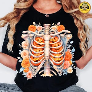 May include: A black t-shirt featuring a graphic print of a human rib cage containing several orange jack-o'-lantern pumpkins, surrounded by a cluster of orange and peach-colored flowers and light blue leaves, centered on the front of the garment.