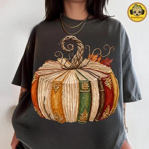 Peut inclure: Un t-shirt gris anthracite arborant un grand motif de citrouille compos&eacute;e de livres anciens empil&eacute;s. La citrouille pr&eacute;sente une tige tress&eacute;e, des d&eacute;tails floraux dor&eacute;s sur les tranches des livres et des feuilles d'automne orange et rouges.