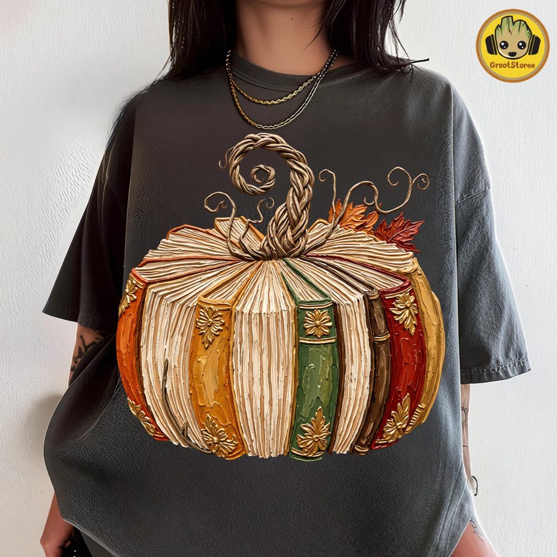 Peut inclure: Un t-shirt gris anthracite arborant un grand motif de citrouille compos&eacute;e de livres anciens empil&eacute;s. La citrouille pr&eacute;sente une tige tress&eacute;e, des d&eacute;tails floraux dor&eacute;s sur les tranches des livres et des feuilles d'automne orange et rouges.