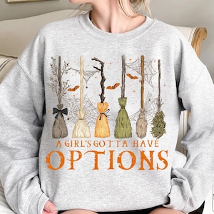 May include: A light gray sweatshirt featuring a Halloween-themed design. The graphic includes several witch brooms, spiderwebs, and bats. The text "A GIRL'S GOTTA HAVE OPTIONS" is printed in orange.