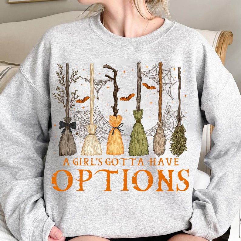 May include: A light gray sweatshirt featuring a Halloween-themed design. The graphic includes several witch brooms, spiderwebs, and bats. The text "A GIRL'S GOTTA HAVE OPTIONS" is printed in orange.