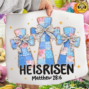 Op de afbeelding: Een wit kussen met drie patchwork kruisen met blauwe en roze bloemen- en ruitpatronen. Elke kruis heeft een decoratieve strik. De tekst "HE IS RISEN" en "Matthew 28:6" is in het zwart gedrukt.